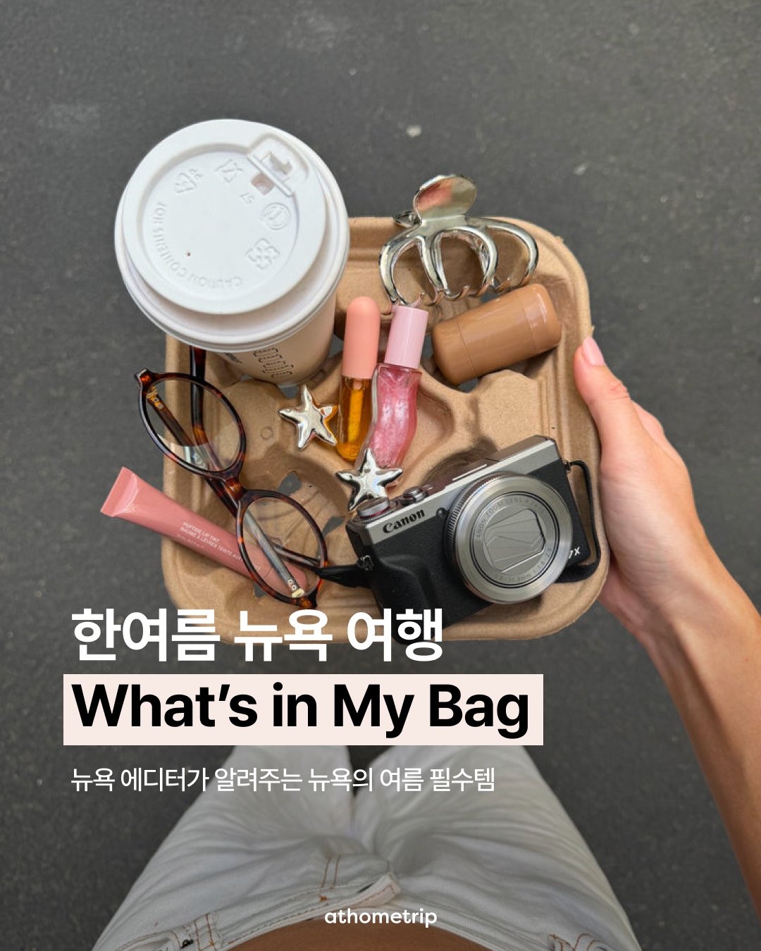 손에 든 종이 트레이 위에 커피잔, 화장품, 카메라, 선글라스가 놓여 있는 실외 장면