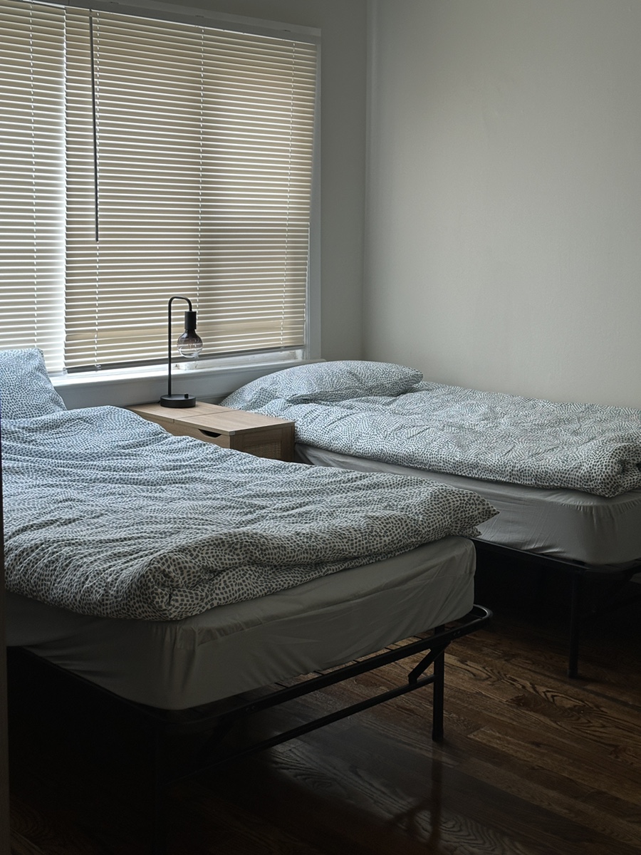 블라인드가 설치된 창문 아래 두 개의 침대와 나무 소재의 테이블이 있는 방