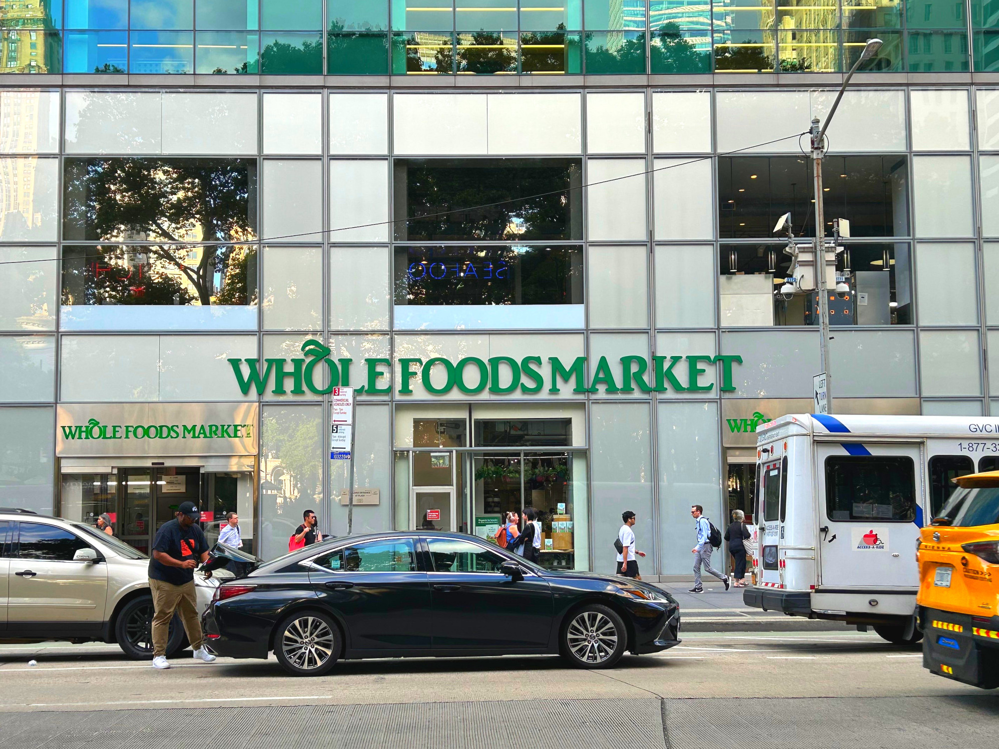 도시 도로 위로 건물과 자동차, 택시가 지나가는 가운데 WHOLE FOODS MARKET 간판이 보이는 장면