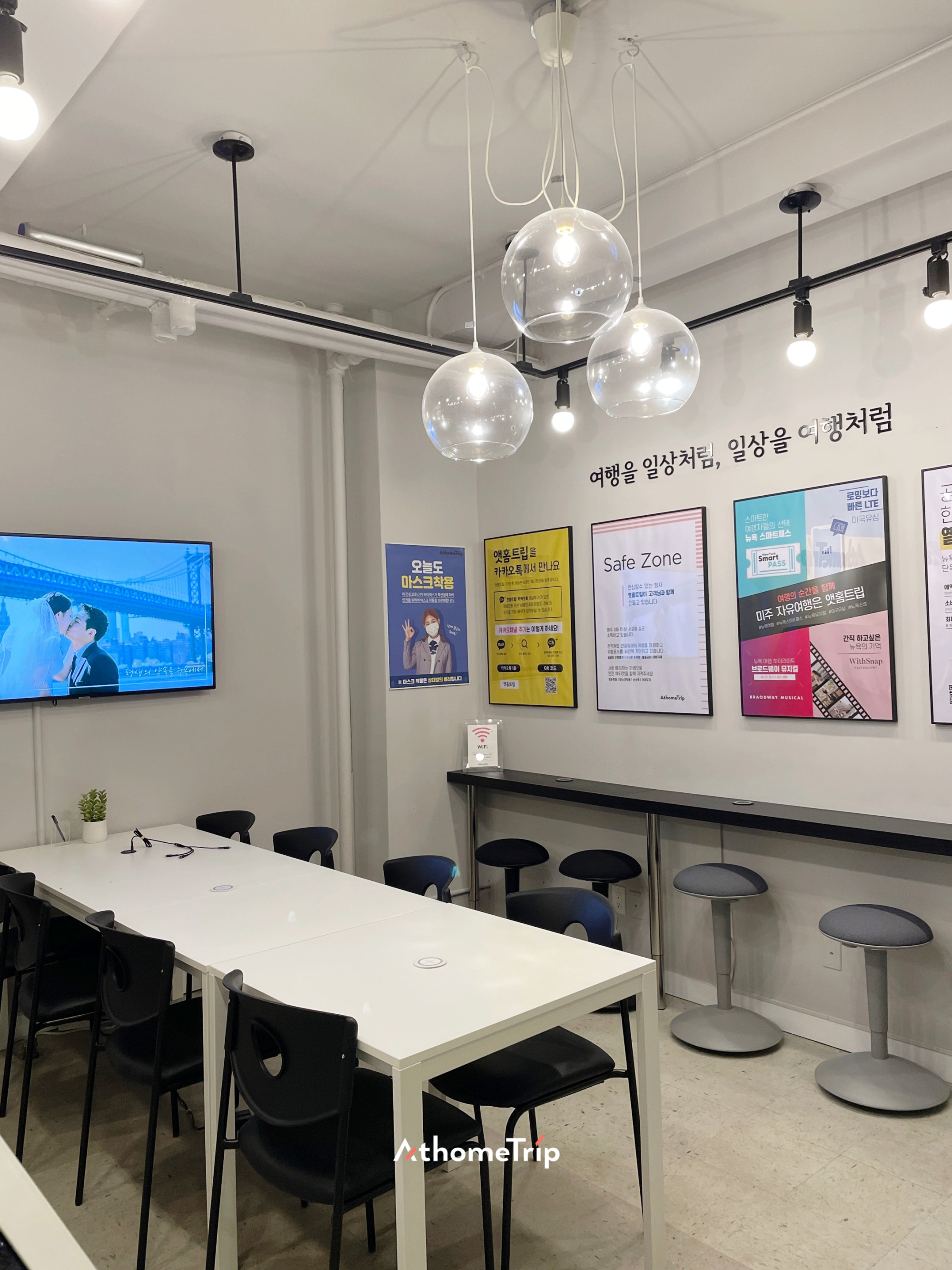 실내 공간에 흰색 테이블, 검은 의자, 벽면의 포스터와 전광판이 보입니다.