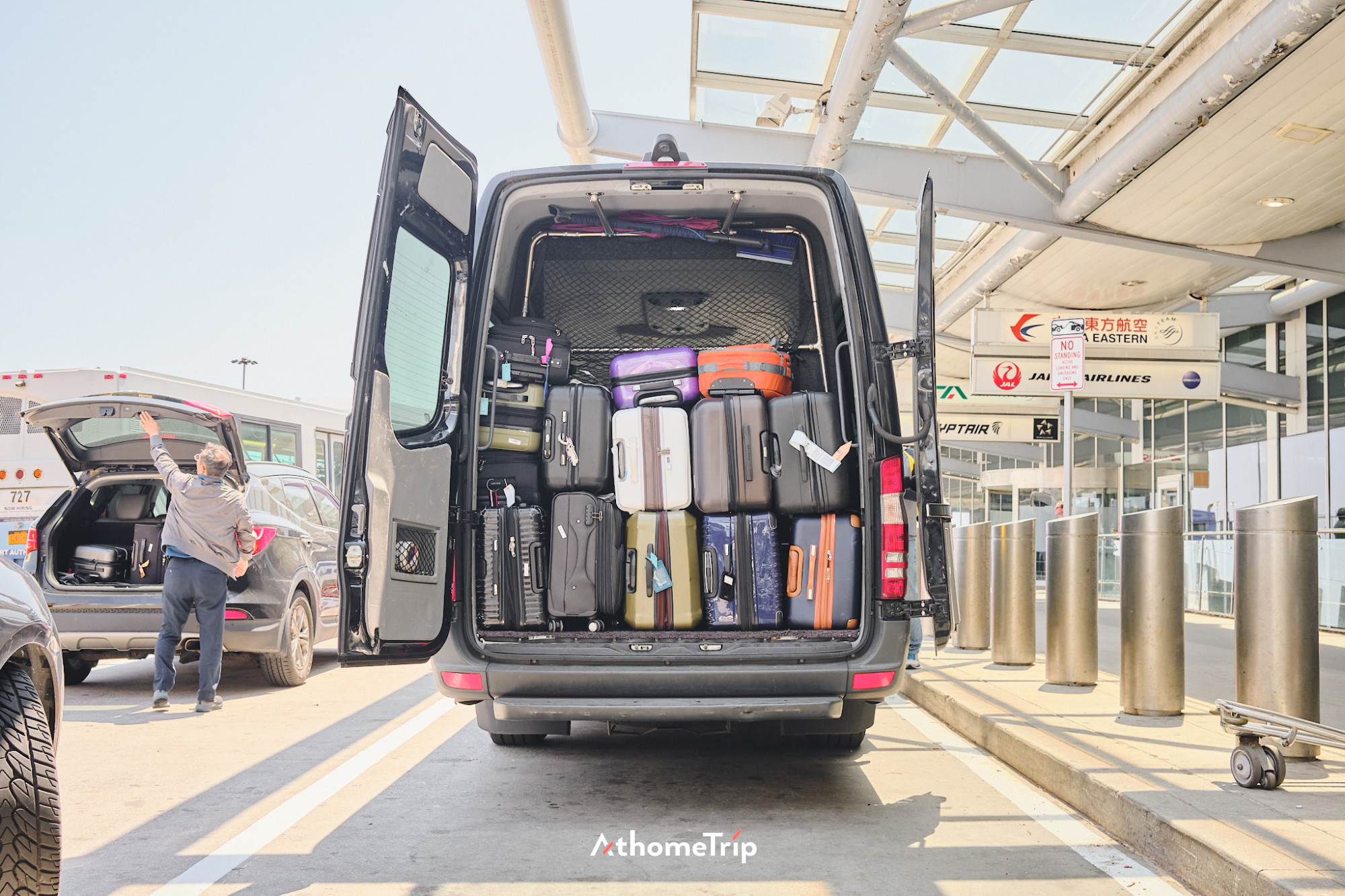 공항 주차장에 세워진 대형 차량의 후면 문이 열려 있으며, 내부에는 여러 개의 여행 가방이 밀집되어 있습니다. 주변에는 다른 차량과 공항 건물이 보입니다.